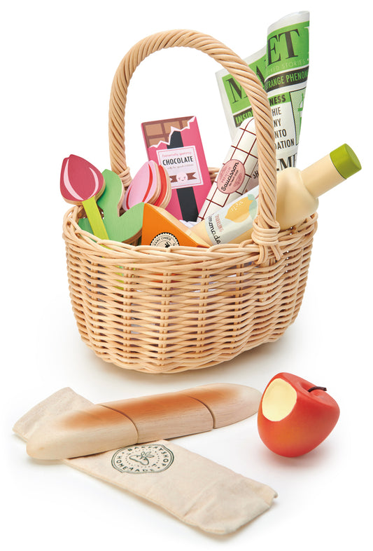 Panier de retour du marché regroupant un ensemble de fruits, légumes et accessoires en bois pour enfant à partir de 3 ans : pomme, baguette et son étui coton, limonade, fromage, chocolat, tulipes, saucissons, journal. Un bel ensemble coloré dans un panier en osier clair. Tender Leaf.