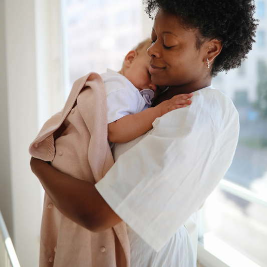 Bébé dans les bras de sa maman, sous une couverture rose en coton biologique rose. Mushie.