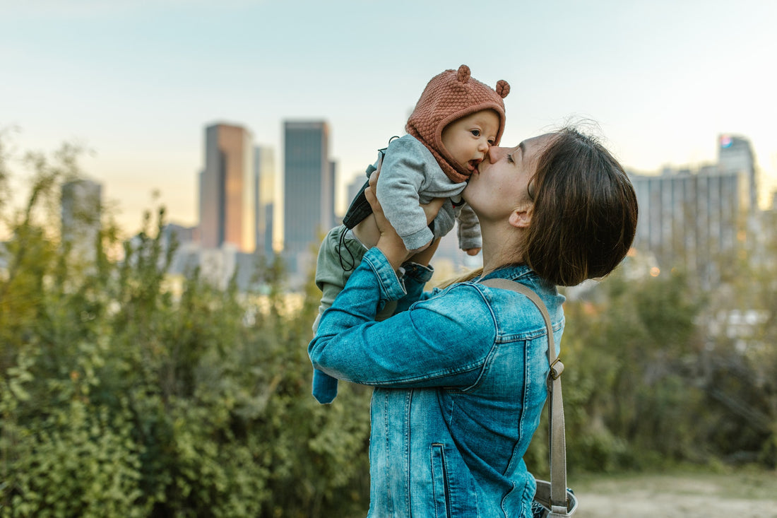 Bien préparer sa promenade avec bébé. Illustration d'une jeune maman embrassant son petit bébé lors d'une sortie, sur fond de quelques arbres et de buildings. 