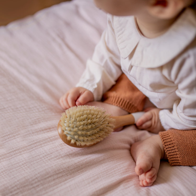 Illustration d'un bébé assis sur un matelas, tenant en sa main une brosse en bois naturel de hêtre Bachca. Idéale pour tous les types de cheveux et le cuir chevelu fragile des nouveau-nés