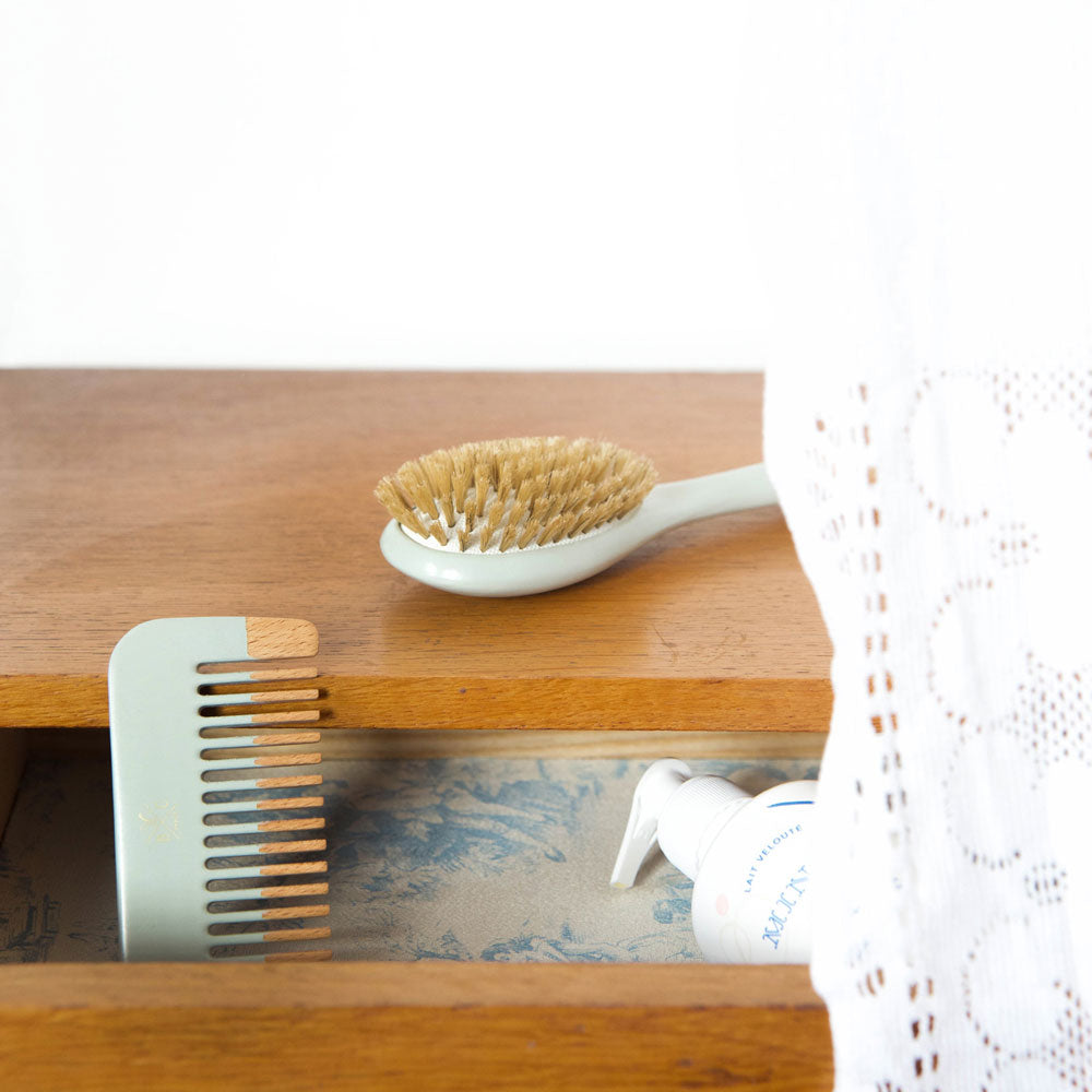 Illustration du contenu du set brosse et peigne pour bébé Bachca, bleu. La brosse en bois pour bébé est posée sur un meuble, et le peigne pour bébé dans le premier tiroir. Bachca Paris.