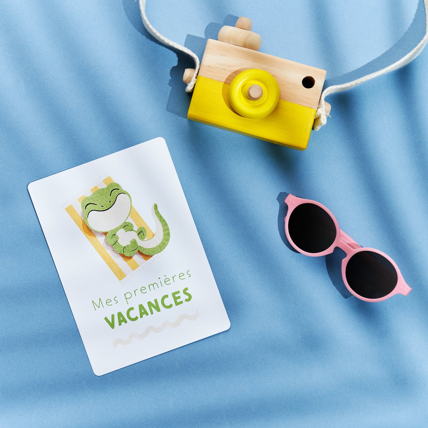 Carte étape première année de bébé : mes premières vacances, carte posée sur un fond bleu avec appareil photo jouet en bois et une paire de lunettes. Illustration petite grenouille tout sourire sur sa serviette de plage à rayures jaune et blanche. Cadeau de naissance premières fois