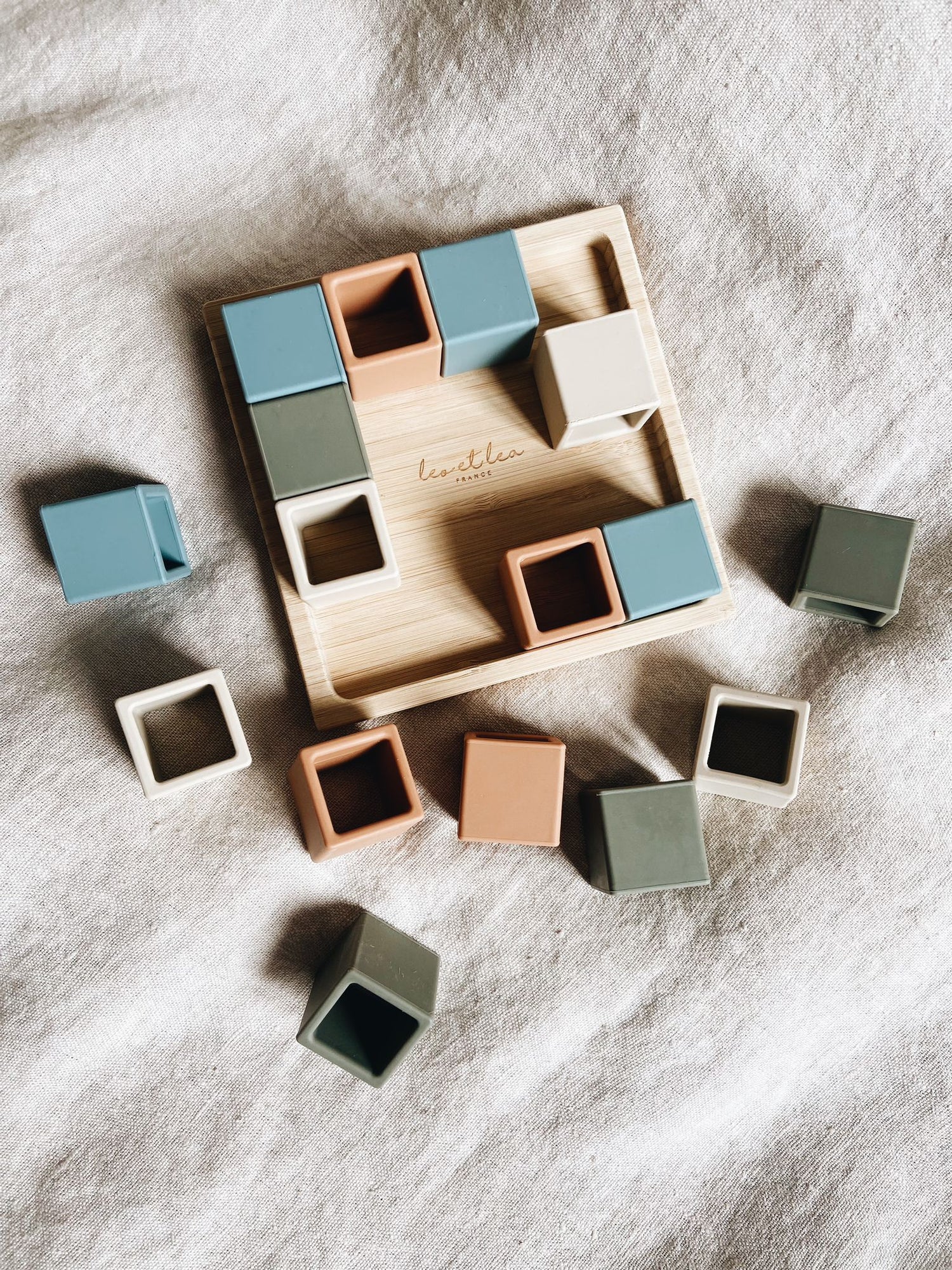 Cubes en silicone de différentes couleurs (bleu, terra cotta, écru, vert sauge) sur plateau en bois. Jeu de construction premier âge. Eveil sensoriel, créativité et imagination.