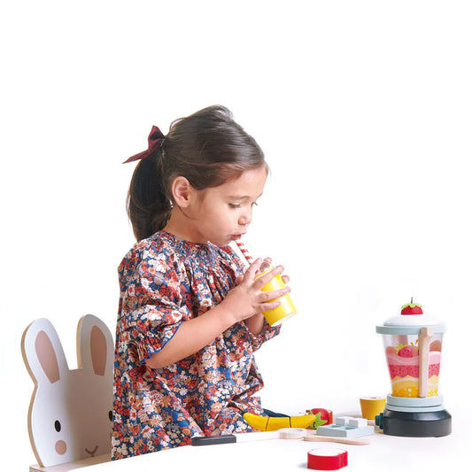 Petite fille brune imitant la dégustation d'un smoothie cuisiné avec son mélanger de fruits en bois. Assise sur une chaise lapin blanche et vêtue d'une robe à fleur estivale. Jouets en bois Tender Leaf respectueux de l'environnement.