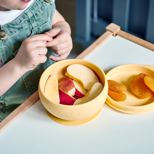 Bol antidérapant couleur jaune miel, en silicone alimentaire. Idéal pour accompagner votre enfant dans ses premiers repas : ici ventousé sur table, avec des quartiers de pomme à déguster par un petit garçon et quelques abricots secs dans le couvercle qui sert de petite assiette.