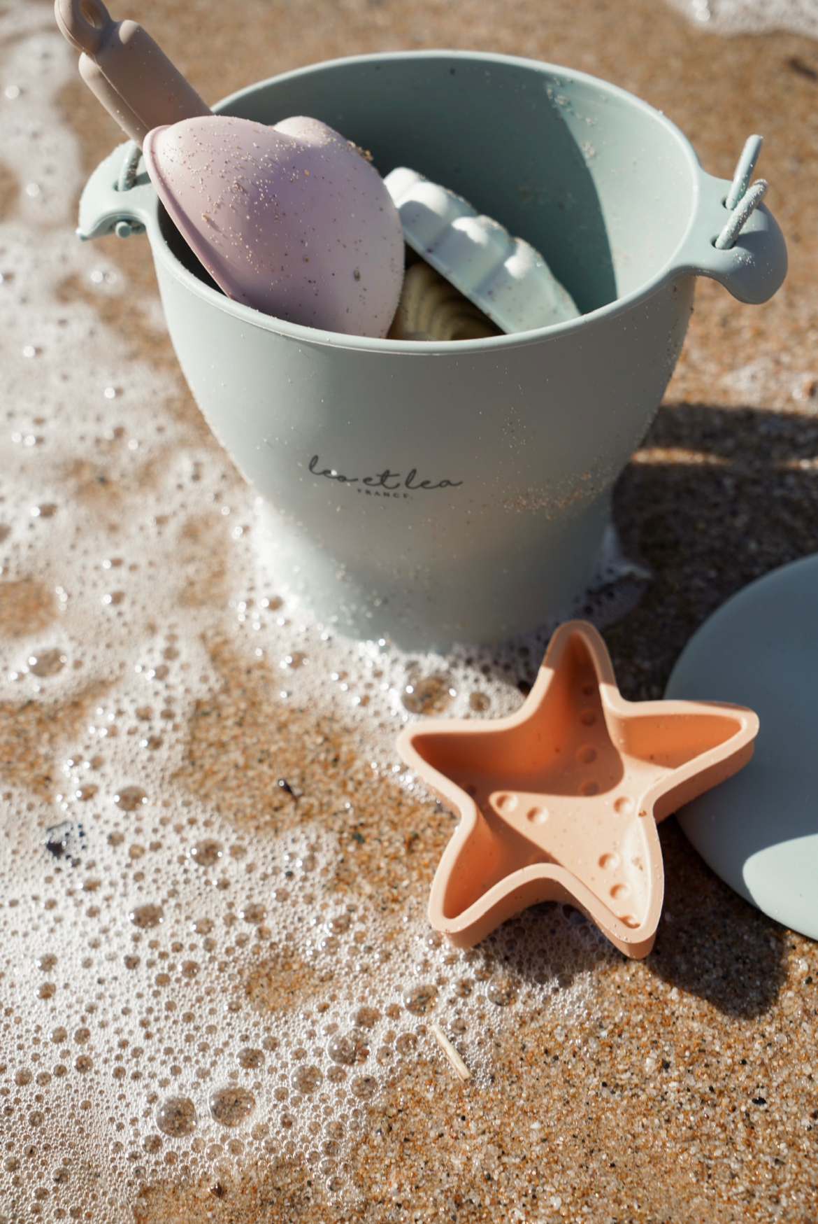 Set de plage mis en situation, posée sur le sable avec l'écume de la mer. Sous le soleil. Un set de jouets idéal pour les premières vacances à la mer. 