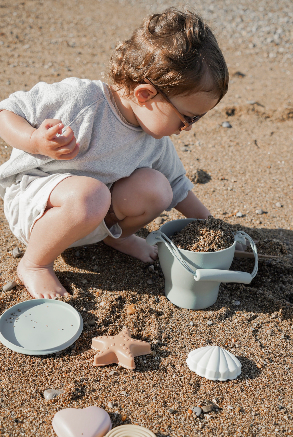 Petit enfant jouant dans le sable avec le set de jouets de plage en silicone Leo et Lea. Premier chateau de sable sous le soleil. Le petit garçon porte un ensemble blanc et gris, et des lunettes de soleil. Ses cheveux bouclés volent au vent. 