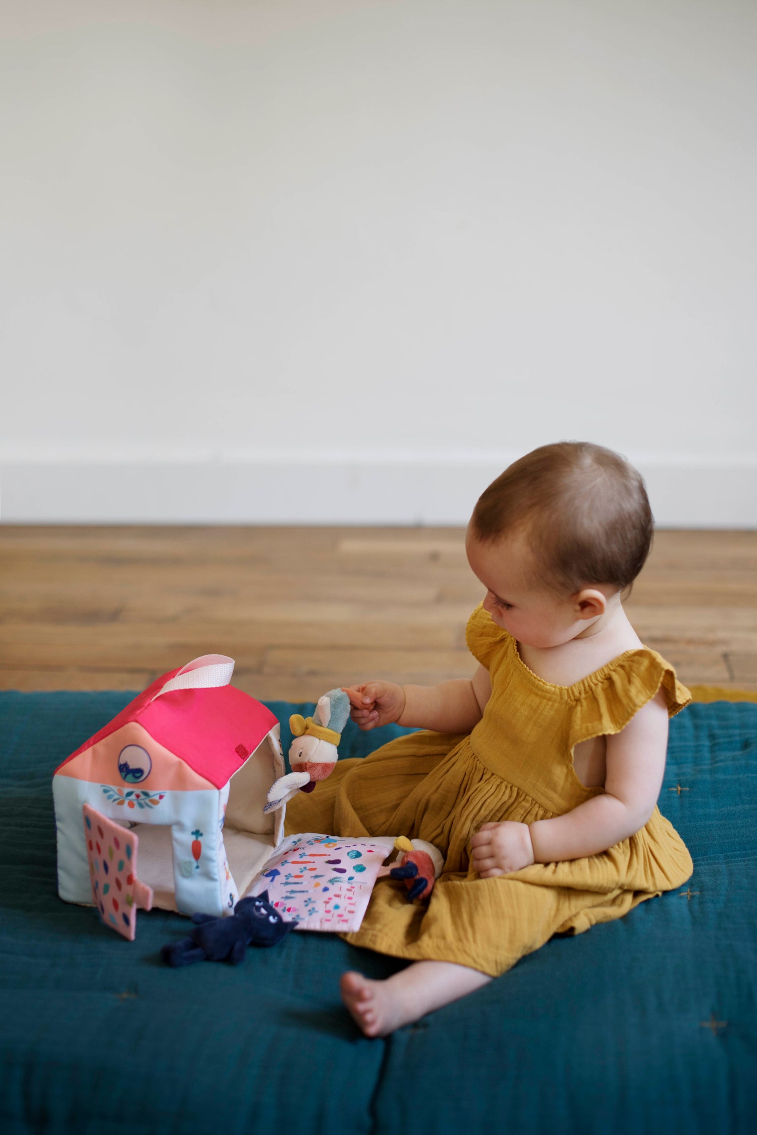 Maison d'activités en tissu pour bébé : sur un tapis d'éveil, une petite fille d'environ 9 mois manipule les trois petites peluches et la maison en tissu de Gabin le Lapin