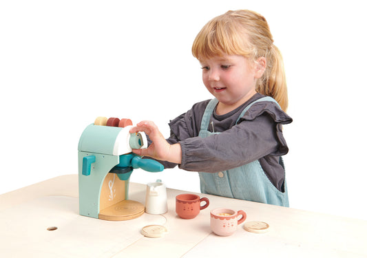 Petite fille jouant à la dinette en bois avec la cafetière en bois Tender Leaf pour faire des bons capuccinos à ses invités. Avec jolis topper pour faire des formes de lait.