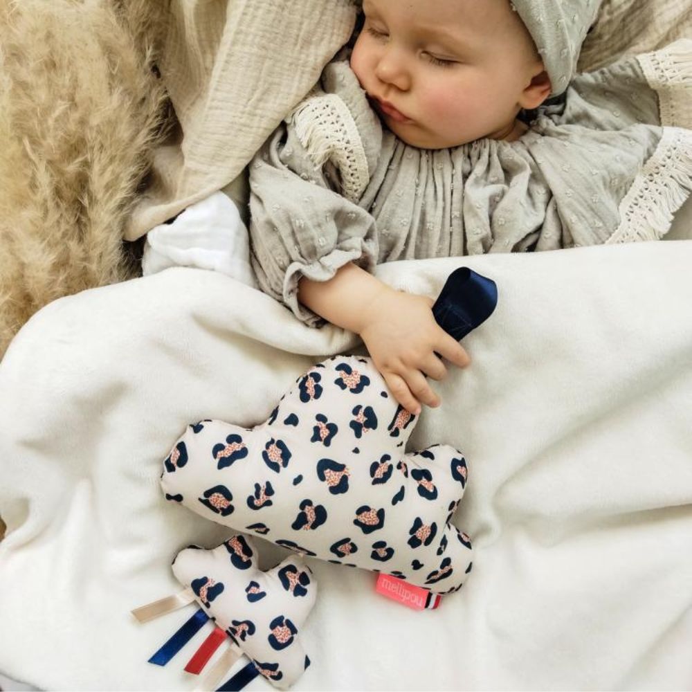 Bébé confortablement installé, endormi dans des draps en gaze de coton et polaire, tenue et petit bonnet beige très clair. Tenant en sa main la peluche musicale beige à tâche léopard bleu conçue par Mellipou et idéale pour s'endormir paisiblement au son d'un berceuse rassurante.