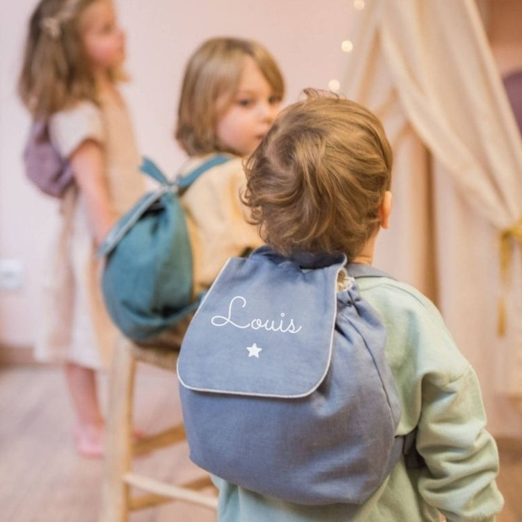 Sac à dos bébé crèche en lin bleu grisé porté par un enfant, personnalisé au prénom : parfait pour la maternelle.