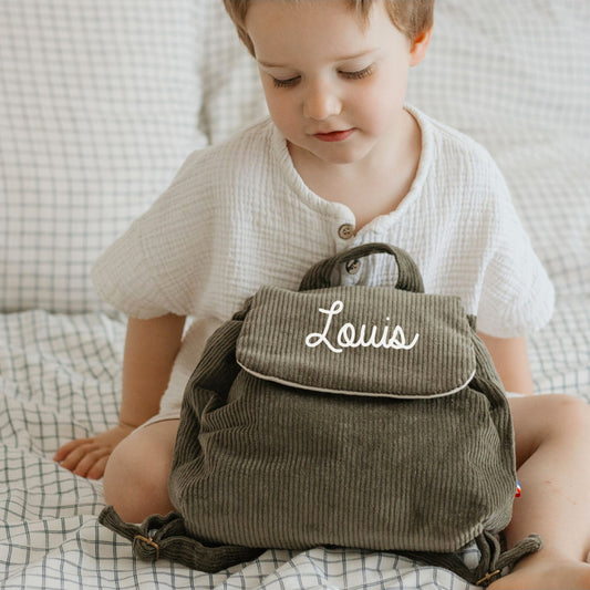 Sac à dos maternelle personnalisé velours cotele kaki posé sur les genoux d’un enfant, format compact pour doudou et goûter.