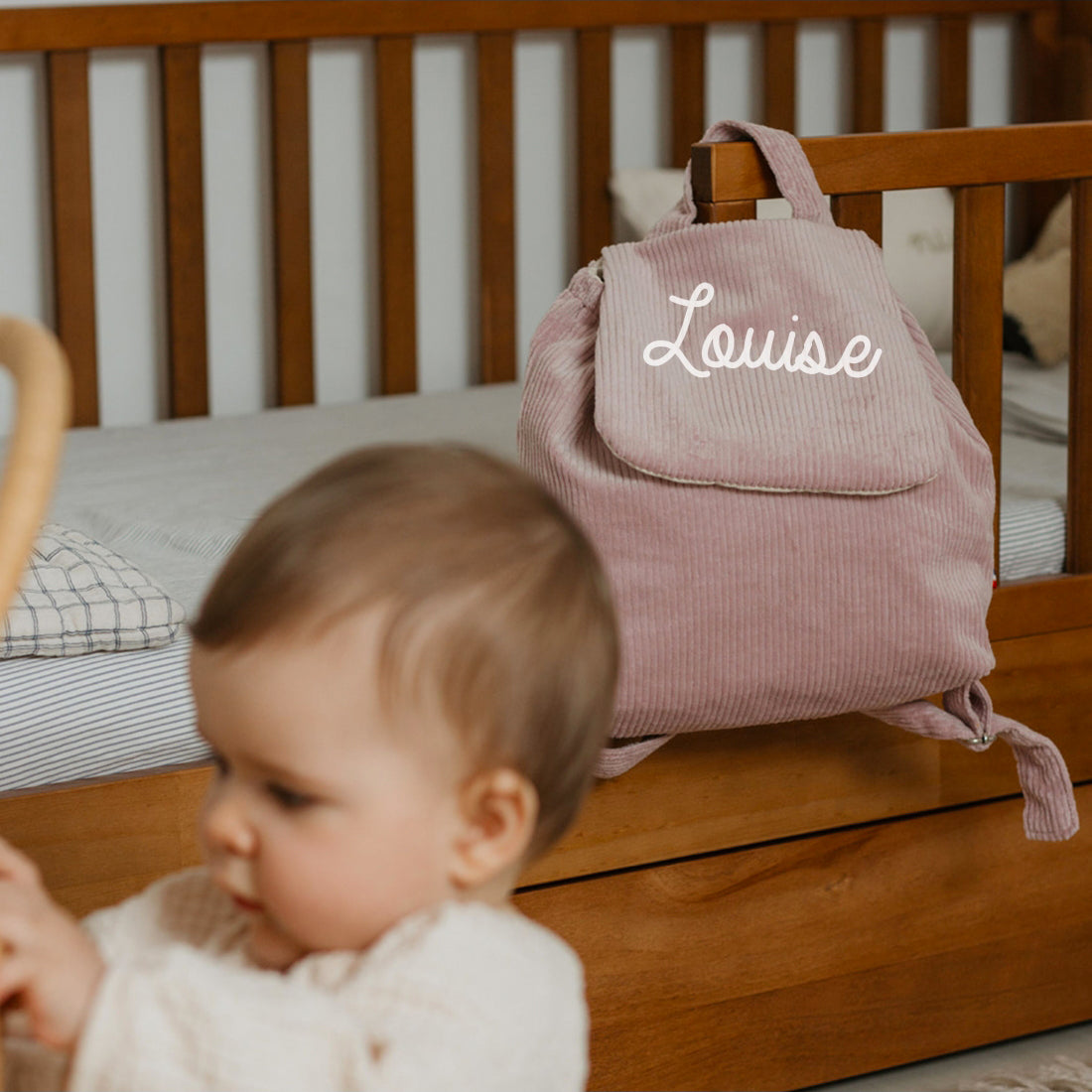 Sac à dos personnalisé en velours côtelé lila, sans motif, posé près du berceau, format idéal doudou et goûter.
