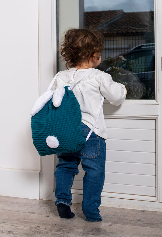 Sac à dos bébé crèche, idéal pour accompagner les aventures de bébé à la crèche ou à l'école. Enfant sur la pointe des pieds regarde par la fenêtre, son petit sac à dos en coton sur le dos (couleur bleu paon). BB&co, marque française d'accessoires textiles pour bébé et enfant.
