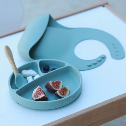 Set repas enfants en silicone alimentaire de la marque Minikoioi. Couleur vert tilleul, mixte. Assiette compartimentée en silicone, cuillère avec manche en bambou, bavoir avec récupérateur de nourriture. Ici posé sur une table, l'assiette antidérapante permet de proposer 3 aliments (figues, myrtilles et coco). Assiette et cuillère bébé vert. 