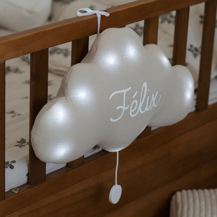 Veilleuse nuage lumineuse beige avec prénom Félix en blanc, suspendue par un ruban au montant d'un lit en bois.