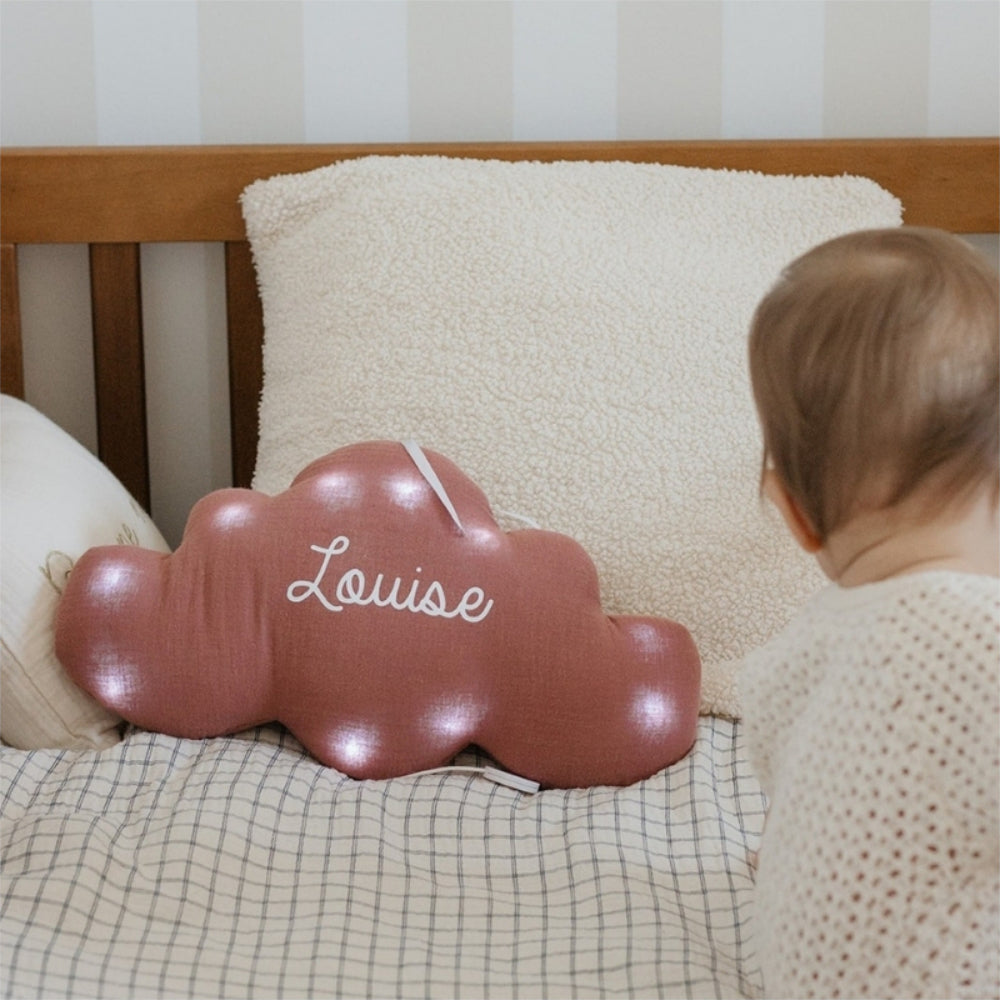 Veilleuse nuage personnalisée en double gaze vieux rose, prénom Louise en blanc et éclairage allumé, photographiée dans une chambre de bébé.