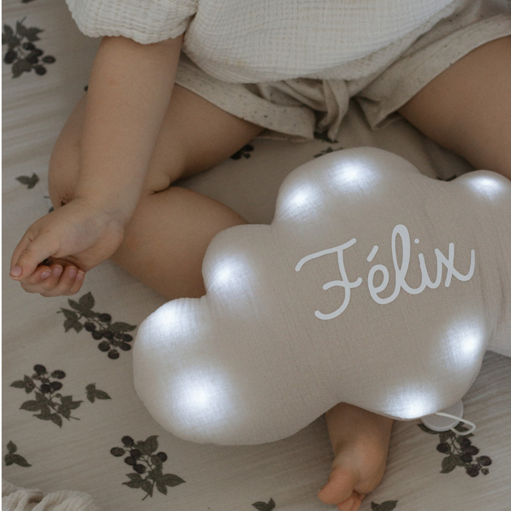 Veilleuse nuage beige allumée et personnalisée au prénom Félix, photographiée à côté d'un bébé habillé de blanc.
