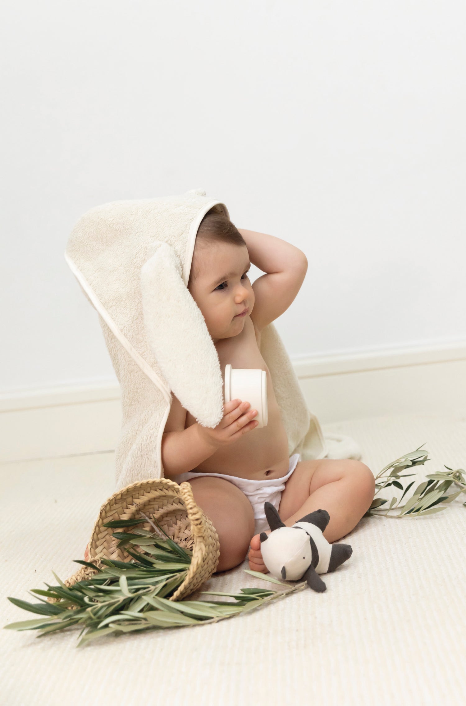 Illustration de la serviette éponge bébé. Un bébé d'environ 8/9 mois avec la cape de bain sur la tête, assis près d'un panier avec feuilles d'Olivier et un petit jouet à la main. Babyshower, made in Europe.