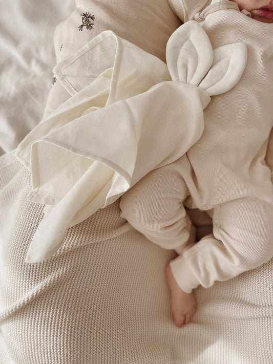 Doudou lange de couleur écru mis en situation, à côté d'un bébé dormant tranquillement sur sa couverture et vêtu d'un body écru