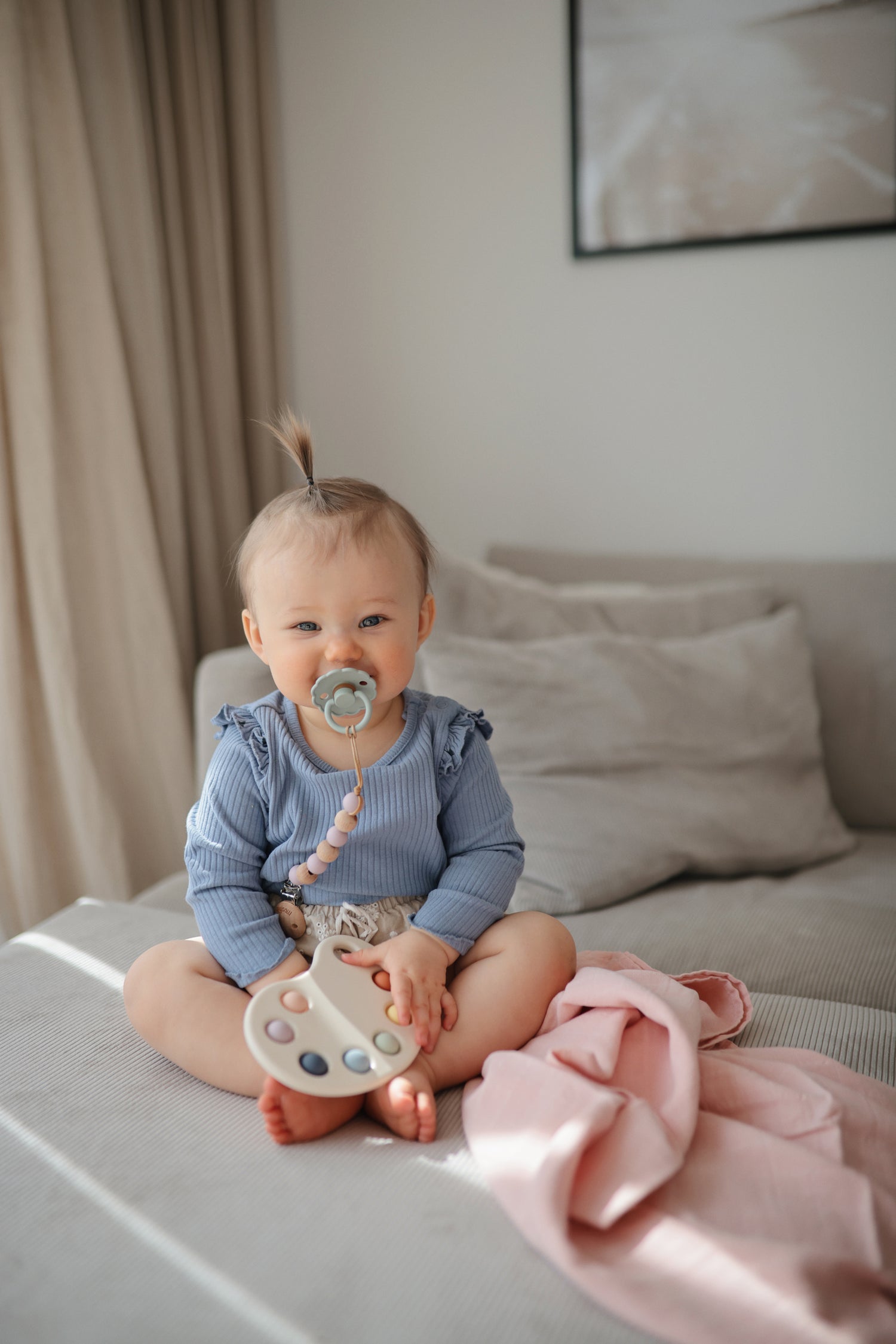 Un bébé joue avec la palette pop it mushie. Installé sur un canapé, vêtu d'un petit top bleu, tétine vert clair en bouche et yeux souriant.