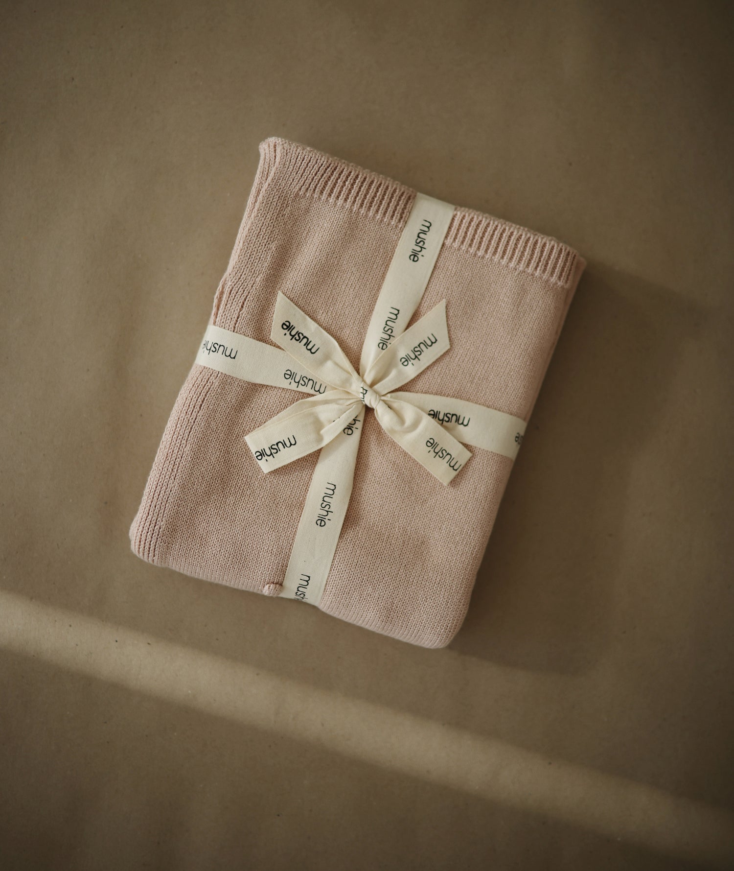 Packaging de la couverture bio pour bébé Mushie, délicatement pliée et entourée d'un ruban marquée Mushie, surmonté d'un noeud. 