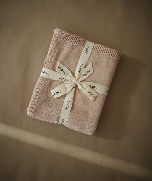 Packaging de la couverture bio pour bébé Mushie, délicatement pliée et entourée d'un ruban marquée Mushie, surmonté d'un noeud. 