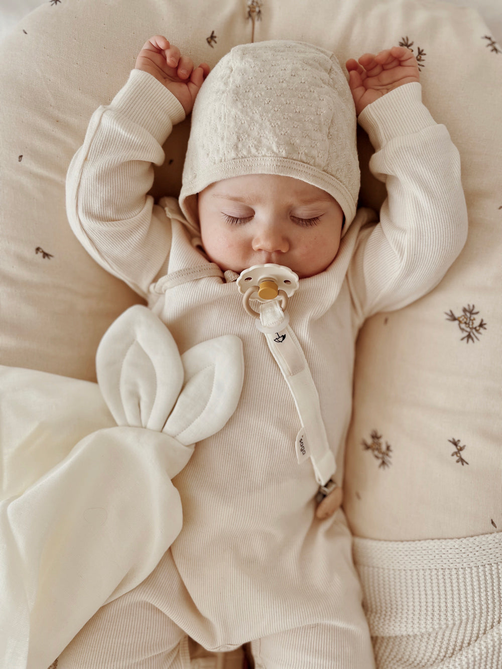 Bébé dormant paisiblement, les bras levés, et à ses côtés un doudou lange lapin Saga écru.